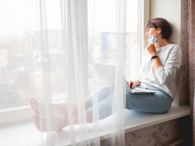 Tıbbi maskeli kadın uzaktan kumanda ile evden çalışıyor. Pencere pervazına dizlerinin üstüne çöküp dizlerinin üstüne çöker. Coronavirüs COVID19 yüzünden karantina altına alındı. Evde yalnızlık.