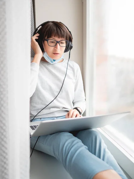 Woman in medical mask remote works from home. She sits on window sill with laptop on knees and headphones. Lockdown quarantine because of coronavirus COVID19. Self isolation at home.