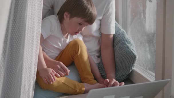 Afgelegen vrouw werkt vanuit huis. Ze zit op de vensterbank met laptop en knappe peuter op haar knieën. Afsluiting quarantaine vanwege het coronavirus COVID19. Zelfisolatie thuis. 10bit V-logL — Stockvideo