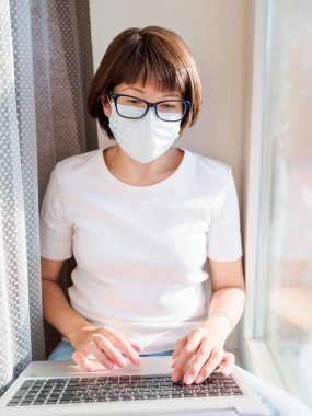 Tıbbi maskeli kadın uzaktan kumanda ile evden çalışıyor. Pencere pervazına dizlerinin üstüne çöküp dizlerinin üstüne çöker. Coronavirüs COVID19 yüzünden karantina altına alındı. Evde yalnızlık.