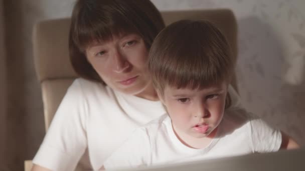 Anne ve küçük oğlu dizüstü bilgisayarla birlikte sandalyede oturuyorlar. Kadın uzaktan kumandayla iş yapmaya çalışıyor ama çocuk oyun ya da çizgi film istiyor. Koronavirüs COVID-19 yüzünden karantinaya alındı. 10bit V günlük kaydı — Stok video