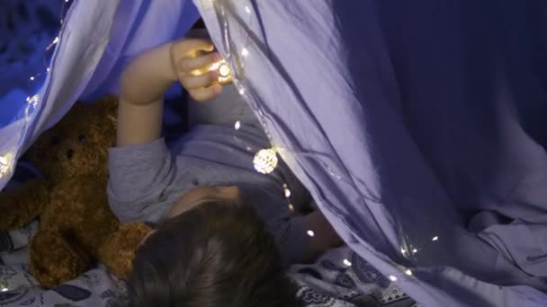 小さな男の子は懐中電灯で読みます。幼児はベッドのリネンシートで作られたテントで遊ぶ。お気に入りの本と居心地の良い夜. — ストック動画
