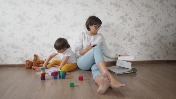 Mutter und Sohn sitzen wegen Coronavirus COVID19 zu Hause in Quarantäne. Mutter arbeitet aus der Ferne mit Smartphone, Sohn spielt mit Spielzeugklötzen. Selbstisolierung zu Hause. — Stockvideo