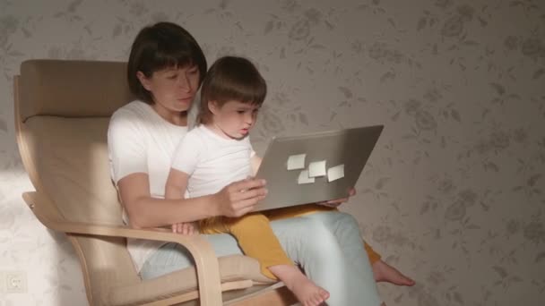 Moeder en haar peuter zitten samen op een stoel met laptop. Vrouw probeert op afstand te werken, maar kind vraagt om spel of tekenfilms. quarantaine afgesloten vanwege het coronavirus COVID-19. 10bit V-logL — Stockvideo