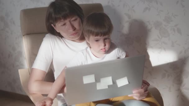 Mor och hennes småbarn sitter tillsammans på en stol med laptop. Kvinnan försöker arbeta på distans, men grabben ber om spel eller tecknade serier. Karantänavstängning på grund av coronavirus COVID-19. 10bit V-logL — Stockvideo