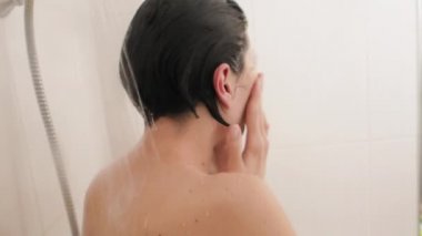Çıplak kadın duş alıyor. Kadın kısa saçlarını suyla yıkıyor. Beyaz banyoda yavaş çekim videosu.