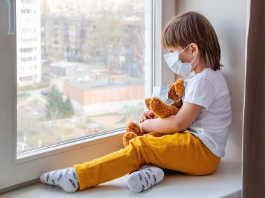 Küçük çocuk tıbbi maskeli oyuncak ayıyla oturuyor. Pelüş oyuncaklı çocuk pencereden dışarı bakıyor. Browh peluş ayı ve çocuk karantinada çünkü Coronavirus COVID-19.
