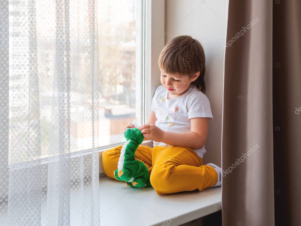 Toddler boy sits with green dragon or dinosaur. Kid with plush toy look through window outside. Child on home quarantine because of coronavirus COVID-19 waiting for somebody.