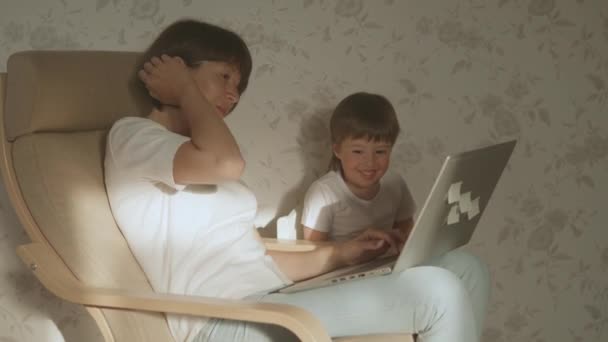 Moeder en haar peuter zitten samen op een stoel met laptop. Vrouw probeert op afstand te werken, maar kind vraagt om spel of tekenfilms. quarantaine afgesloten vanwege het coronavirus COVID-19. 10bit V-logL — Stockvideo