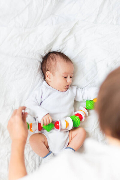 Woman plays with newborn boy. Baby's first toy - colorful rattle garland. Top view on little child in white onesie. Morning bedtime in cozy home.