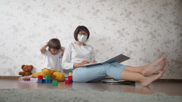 Mamma och son sitter i karantän på grund av coronavirus COVID19. Mor jobbar på distans med laptop, son leker med leksaksblock. Självisolering i hemmet. — Stockvideo