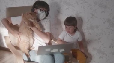 Anne, küçük çocuk ve kedi dizüstü bilgisayarla birlikte sandalyede oturuyorlar. Kadın uzaktan kumandayla iş yapmaya çalışıyor ama çocuk oyun ya da çizgi film istiyor. Koronavirüs COVID-19 yüzünden karantinaya alındı. 10bit V günlük kaydı