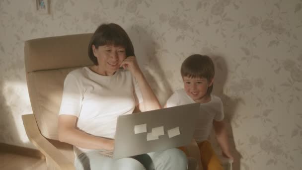 Madre y su niño pequeño se sientan juntos en la silla con el ordenador portátil. Mujer trata de trabajo remoto, pero el niño está pidiendo juego o dibujos animados. bloqueo de cuarentena por coronavirus COVID-19. V-logL de 10bit — Vídeo de stock