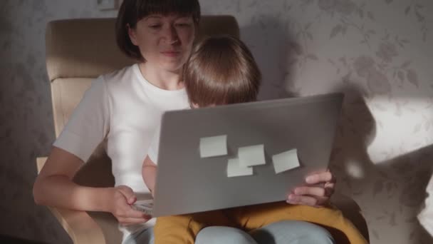 Moeder en haar peuter zitten samen op een stoel met laptop. Vrouw probeert op afstand te werken, maar kind vraagt om spel of tekenfilms. quarantaine afgesloten vanwege het coronavirus COVID-19. 10bit V-logL — Stockvideo