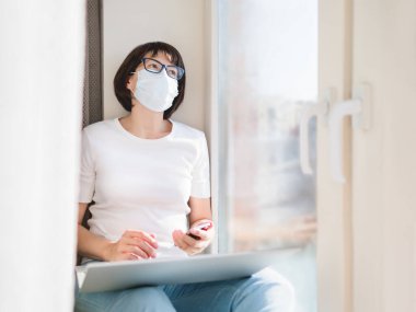 Tıbbi koruyucu maskeli kadın evden uzakta çalışıyor. Akıllı telefon ve dizüstü bilgisayarla pencere eşiğinde oturuyor. Coronavirüs COVID19 yüzünden karantinaya alınmıştır.. 