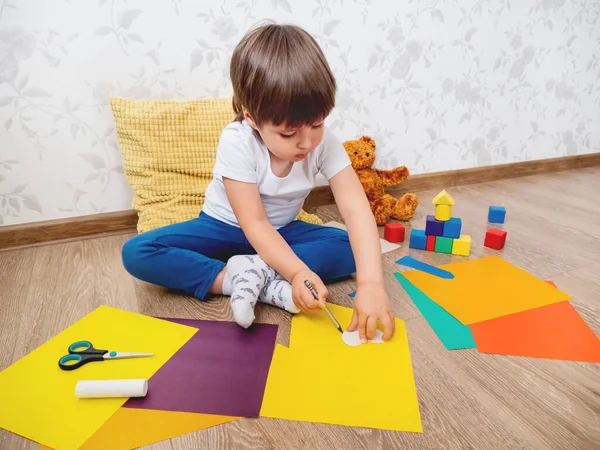 Toddler boy learns to cut colored paper with scissors. Kid sits on floor in kids room with toy blocks and teddy bear. Educational classes for children. Developing feeling sensations and fine motor skills at home.