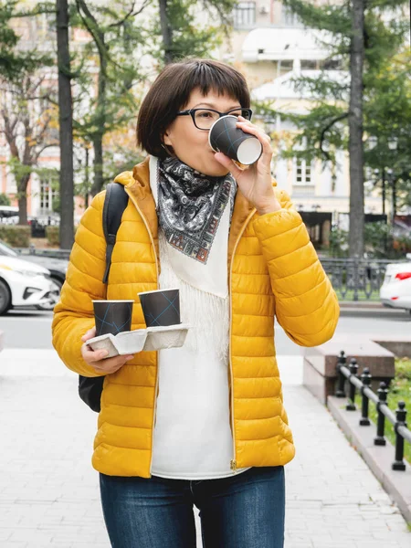 Take away coffee. Women in bright yellow jacket holds paper cup with freshly brewed cappuccino. Hot beverage on cool autumn day.