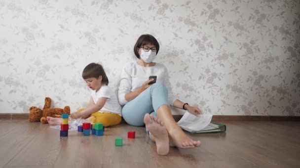 Mamá e hijo se sientan en cuarentena a causa del coronavirus COVID19. Madre trabaja a distancia con el teléfono inteligente, hijo juega con bloques de juguetes. Autoaislamiento en casa . — Vídeos de Stock