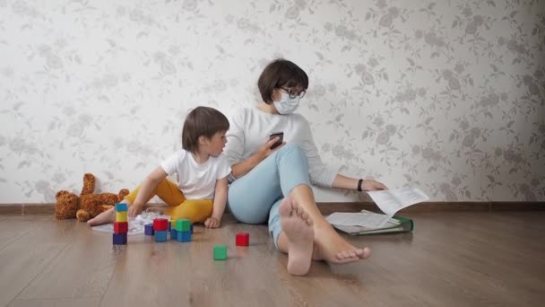 Mutter und Sohn sitzen wegen Coronavirus COVID19 zu Hause in Quarantäne. Mutter sitzt mit Smartphone, Sohn spielt mit Spielzeugklötzen. Selbstisolierung zu Hause. — Stockvideo