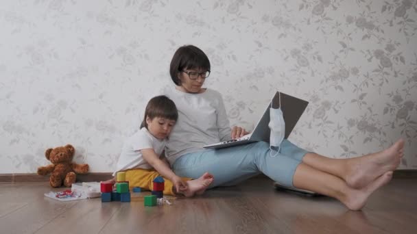 Mama i syn siedzą w domowej kwarantannie z powodu koronawirusu COVID19. Matka zdalnie pracuje z laptopem, syn bawi się klockami. Samoizolacja w domu. — Wideo stockowe