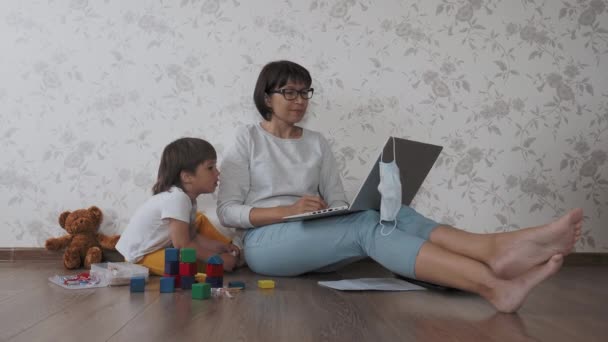 Мама и сын сидят дома в карантине из-за коронавируса COVID19. Мать работает с ноутбуком, сын играет с игрушечными блоками. Самоизоляция дома . — стоковое видео