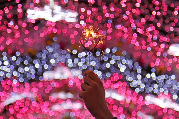 Mão segurando sparkler na forma de um coração no fundo de luzes coloridas desfocadas — Fotografia de Stock