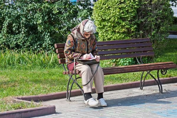 La anciana se sienta en el banco y desentraña un crucigrama en el parque de Volgogrado —  Fotos de Stock