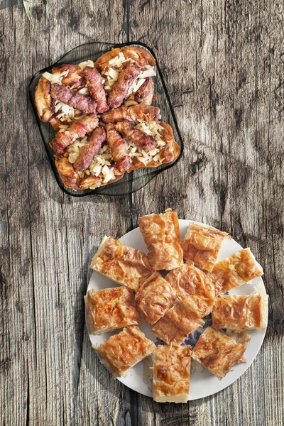 Plateful Of Grilled Minced Meat Loafs Cevapcici Rolled In Bacon With Chicken Meat and Gibanica Crumpled Cheese Pie Set On Old Knotted Cracked Wood — стоковое фото