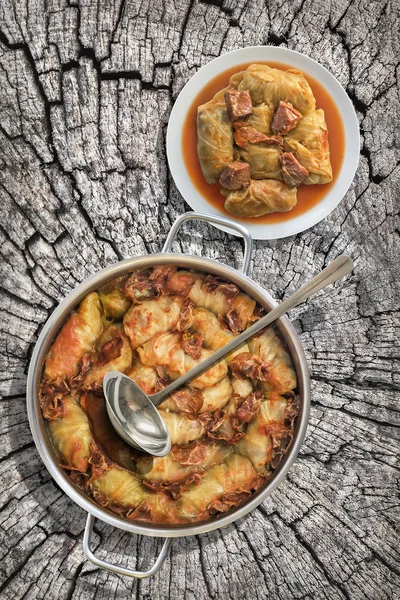 Plateful de rollos de col rellenos con carne picada cocida en salsa de tomate con rebanadas de jamón ahumado en olla de acero inoxidable conjunto en viejo tocón agrietado — Foto de Stock