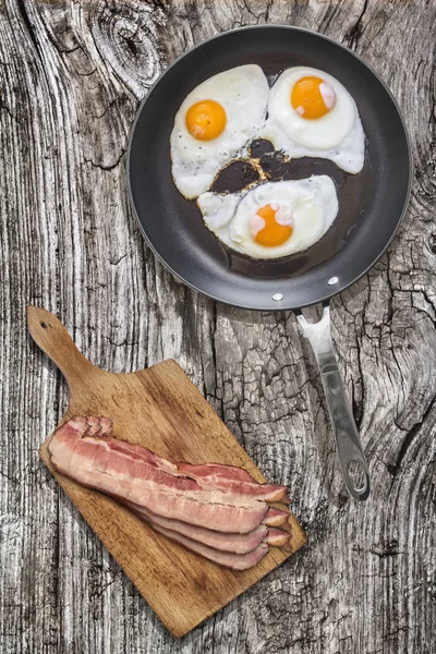 Ηλιόλουστη πλευρά προς τα πάνω τηγανιτά αυγά σε τηγάνι βαρέως τύπου με τεφλόν με χοιρινό μπέικον σε ξύλο κοπής ορίστε την παλιά ραγισμένη ξύλινο τραπέζι πικνίκ — Φωτογραφία Αρχείου