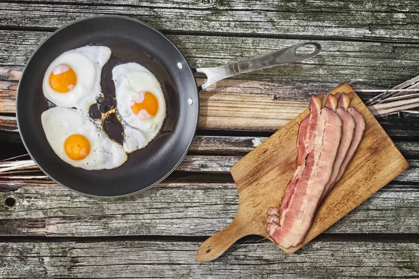 Sunny Side Up Fried Eggs In Heavy Duty Teflon Frying Pan With Pork Bacon Rashers On Cutting Board Set On Old Cracked Wooden Picnic Table
