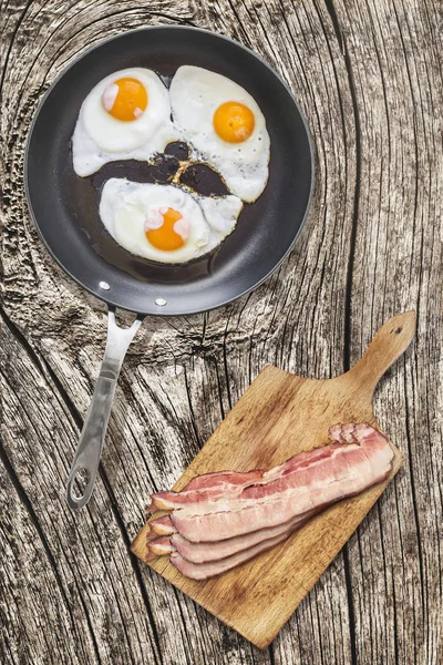 Ağır hizmet Teflon tavada domuz pastırma Rashers üzerinde kesme tahtası ile kızarmış yumurta kadar güneşli tarafı eski kırık ahşap piknik masa ayarla — Stok fotoğraf