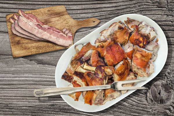 Plateful of Spit Roasted Pork Shoulder Slices and Bacon Rashers on Cutting Board Set on Old Rustic Knotted Wooden Picnic Table