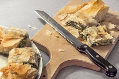Fırından yeni çıkmış ahşap kesme tahtası üzerinde tırtıklı bıçakla dilimlenmiş geleneksel Sırp peynirli ıspanaklı rulo pasta ve porselen tabakta servis