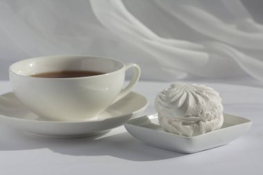 Küçük bir tabakta bir lokum ve arka planda peçesi olan beyaz bir tabakta beyaz bir fincan siyah çay. Doğal gün ışığı.