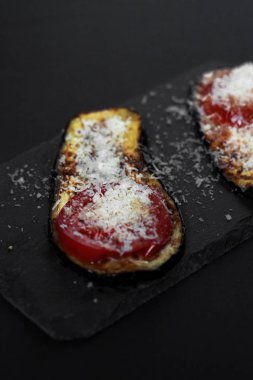 Siyah zemin üzerinde domates ve peynirli ızgara patlıcan dilimleri. Kapat..