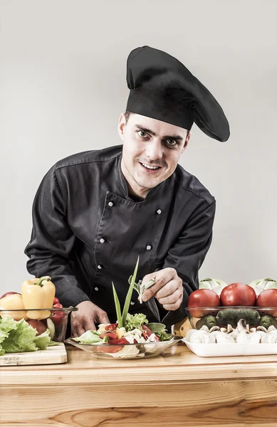 ブルネットハンサムな男 料理人は黒の制服を着用し 木製のテーブルの上にキッチンで野菜を調理 — ストック写真
