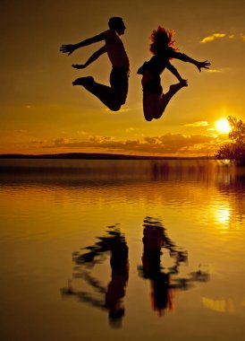 Bir çift - erkek ve kadın sahilde günbatımı bulutlu gökyüzü turuncu güneş arka plan ile sarı su yansıması ile atlama silüeti