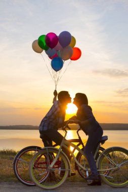 iki kişi birbirine bakarak romantik tarihte. Bisiklet üzerinde oturan genç bir çift. günbatımı bulutlu gökyüzü arka plan üzerinde renkli balonlar tutan adam. 
