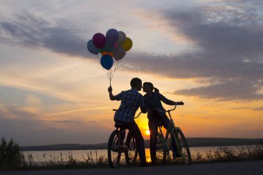 iki kişi birbirine bakarak romantik tarihte. Bisiklet üzerinde oturan genç bir çift. günbatımı bulutlu gökyüzü arka plan üzerinde renkli balonlar tutan adam. 