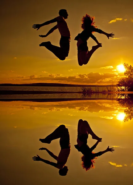 黄色の水の反射と背景がオレンジ色の太陽の日没の曇り空のビーチでジャンプ男女のカップルのシルエット — ストック写真