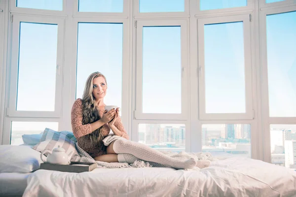 Young beautiful  woman sitting home on the bed by the window. Lazy day off concept. Curly young girl  drinking coffee or tea in bedroom