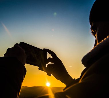 kış dağlarının üzerinde gün batımında cep telefonu tutan adam ellerin silueti. erkek telefonunuzda bir fotoğraf yapma. Güneşin parıltısı silueti aydınlatır.. 