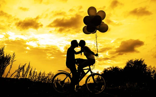 Silhouettes of two beautiful adult people  sitting at  bicycle.  Girl holding colorful balloons at sunset