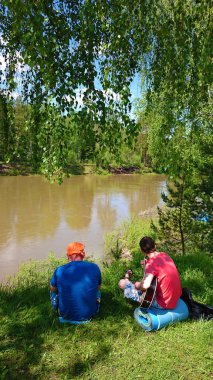 macera, seyahat, turizm ve insan kavramı - akustik gitar ile iki adam geri görünümü ve Rusya'da yaz ormanına karşı kamp nehir bakıyor