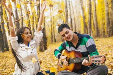 Genç Latin asıllı şirin kız ve arka planda sarı sonbahar orman uçan ile akustik gitar çalmak adam portresini bırakır.