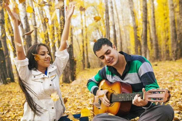 Genç Latin asıllı şirin kız ve arka planda sarı sonbahar orman uçan ile akustik gitar çalmak adam portresini bırakır.