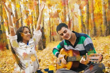 Genç Latin asıllı şirin kız ve arka planda sarı sonbahar orman uçan ile akustik gitar çalmak adam portresini bırakır.