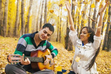 Genç Latin asıllı şirin kız ve arka planda sarı sonbahar orman uçan ile akustik gitar çalmak adam portresini bırakır.