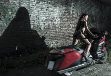 Genç redhead motorcu kadın karanlık gece kırmızı tuğla duvar arka plan üzerinde kırmızı modern motosiklet.